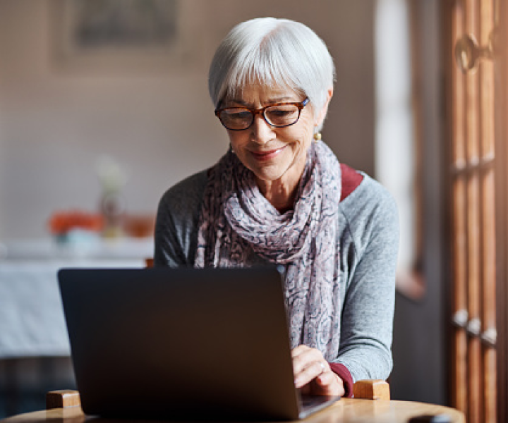 A mature woman in a gray scarf sitting at laptop using Frontier Lifeline Services, a program to help make communications services more affordable for low-income consumers